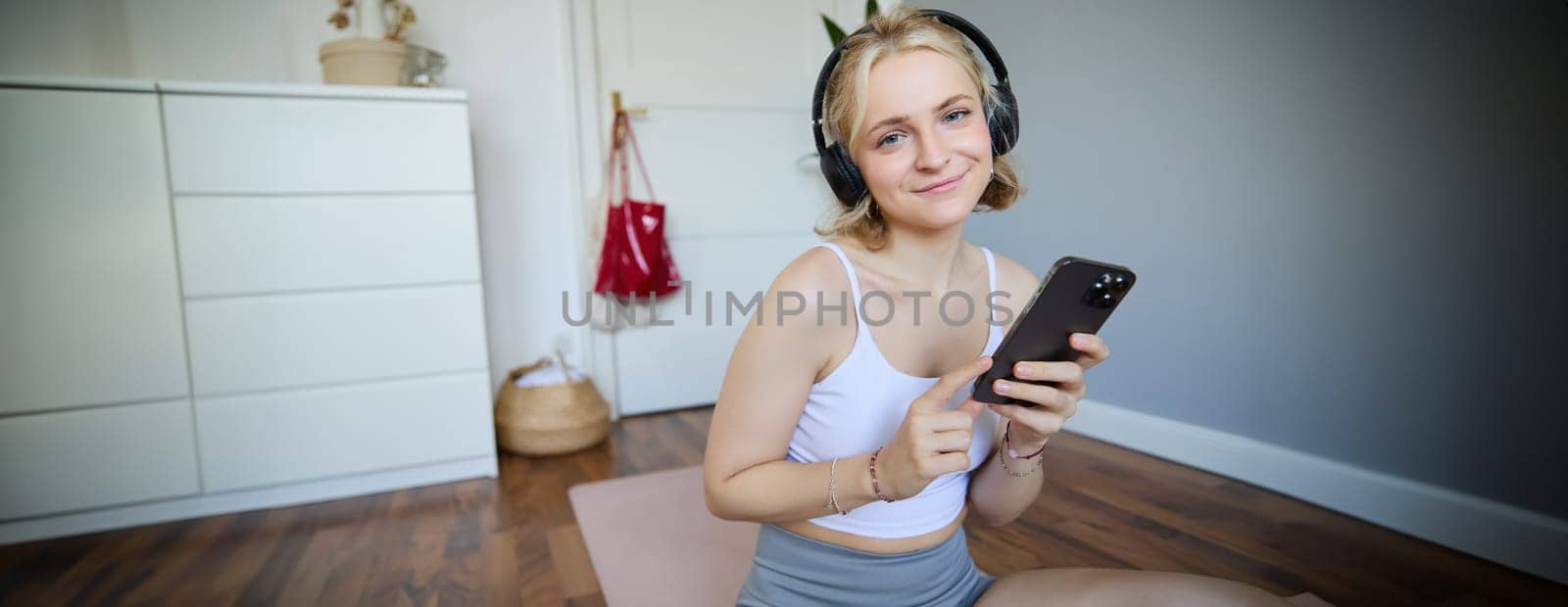 Portrait of smiling fitness woman, wearing wireless headphones, holding mobile phone, working out with sports app on smartphone, sitting on yoga rubber mat, exercising at home by Benzoix