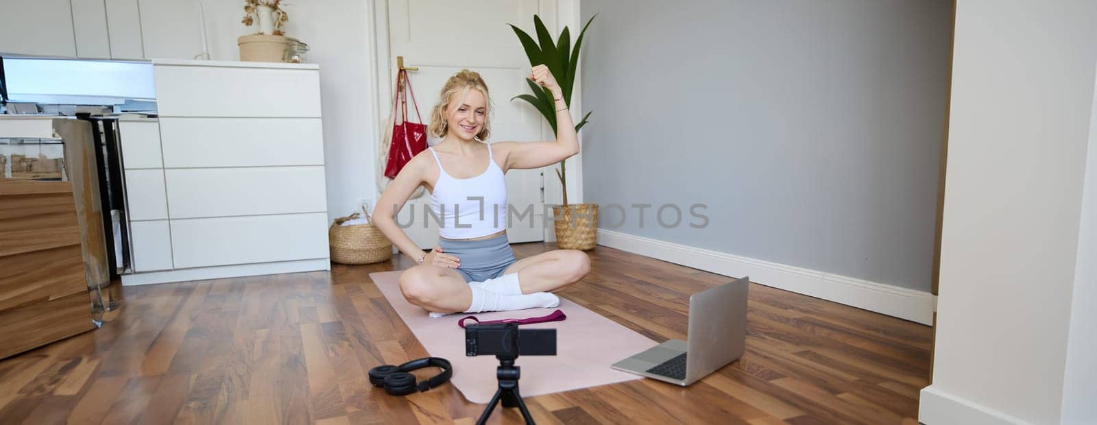 Portrait of young female athlete, fitness trainer recording vlog, training session on digital camera, sitting in a room on rubber yoga mat, showing exercises by Benzoix