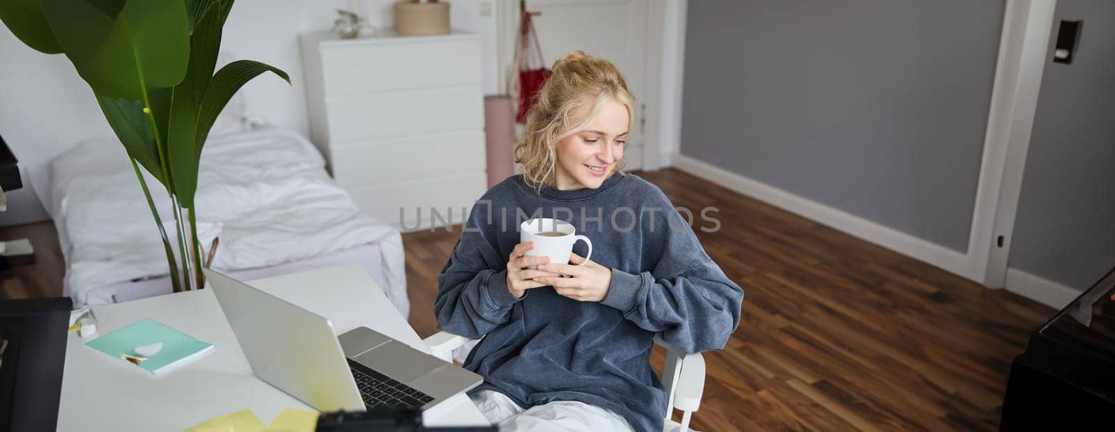 Portrait of young smiling woman, vlogger recording lifestyle video in her room, sits in front of laptop and digital camera, drinks tea, creates content for social media account.