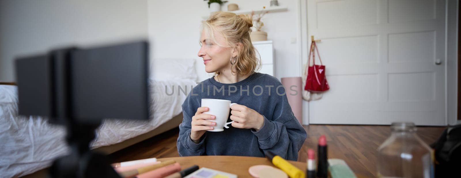 Image of young woman, makeup vlogger, sitting in bedroom with digital camera, drinking tea and talking, creating lifestyle video, social media content.