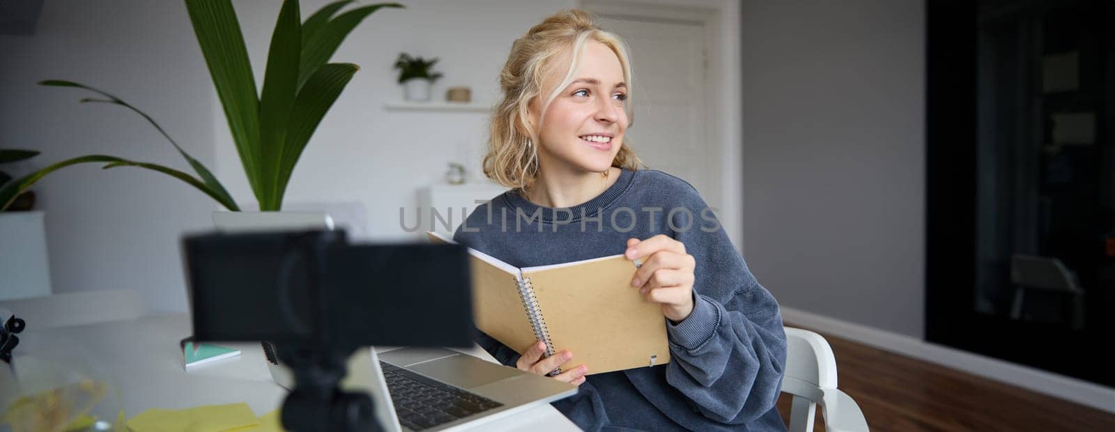 Portrait of smiling blond woman, sitting in bedroom, using laptop and digital camera, recording video for lifestyle blog, reading, using her notebook.