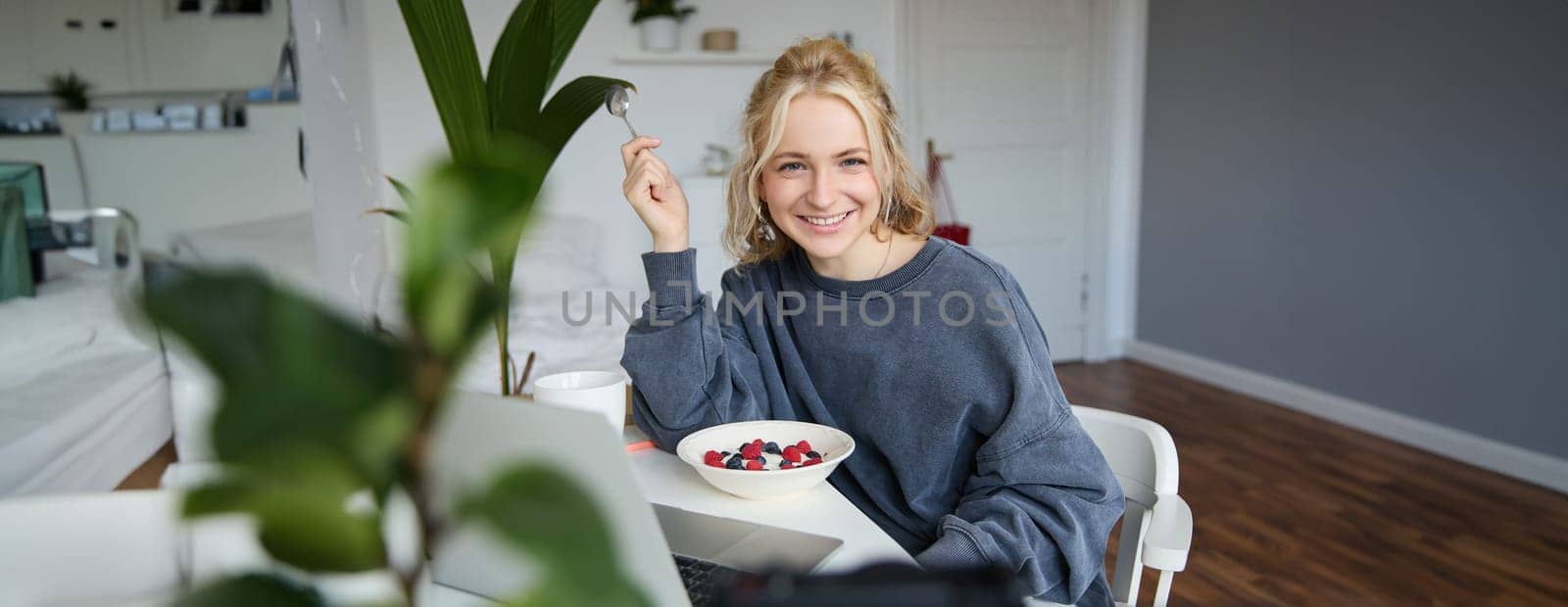 Portrait of young smiling woman, sitting in room, watching videos on laptop, eating lunch or breakfast, healthy meal.