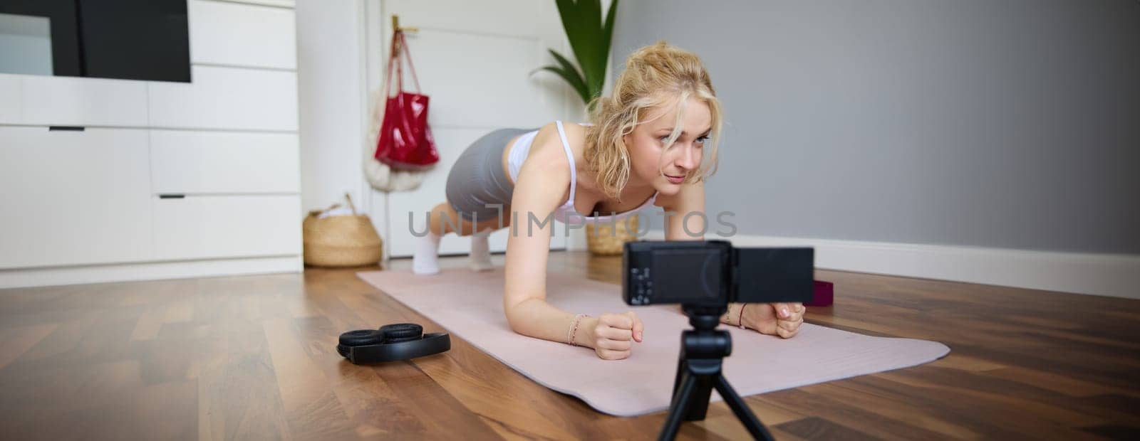 Young woman, personal fitness instructor records video of herself standing in a plank, using rubber yoga mat and digital camera at home.