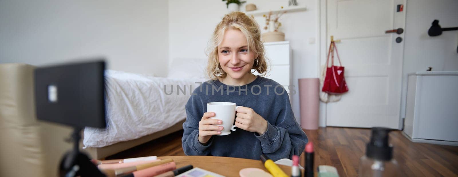 Image of young woman, makeup vlogger, sitting in bedroom with digital camera, drinking tea and talking, creating lifestyle video, social media content by Benzoix