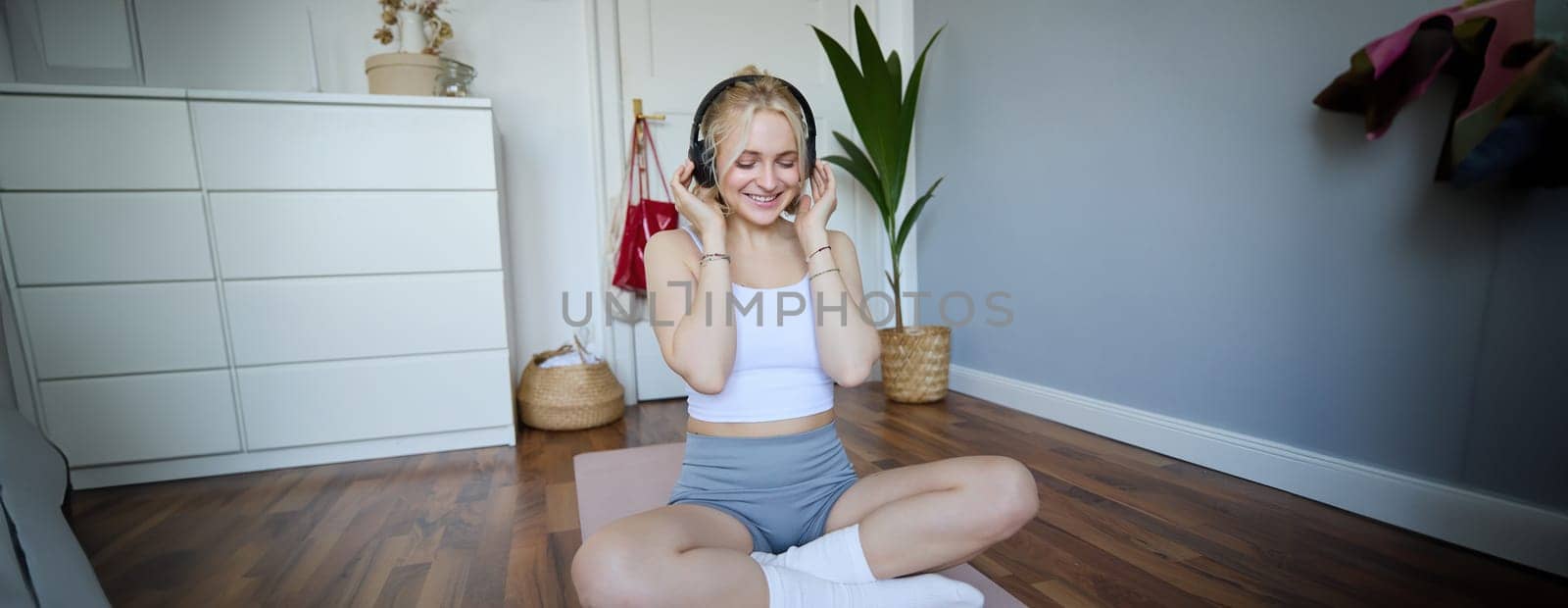 Portrait of happy, beautiful athletic woman, working out at home, listening music to boost energy while active training session, using wireless headphones and rubber yoga mat by Benzoix