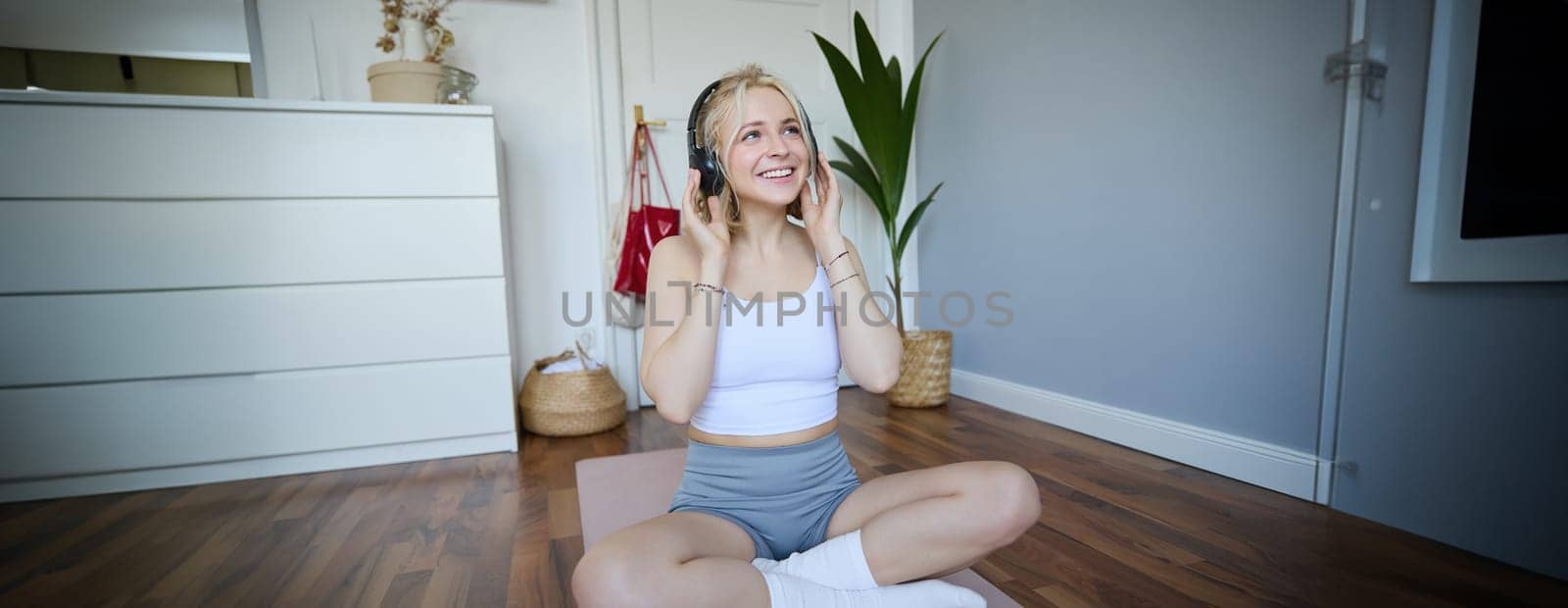 Sport and wellbeing concept. Smiling woman in wireless headphones, sitting on yoga mat, listens to music and workout at home, enjoys fitness training session.