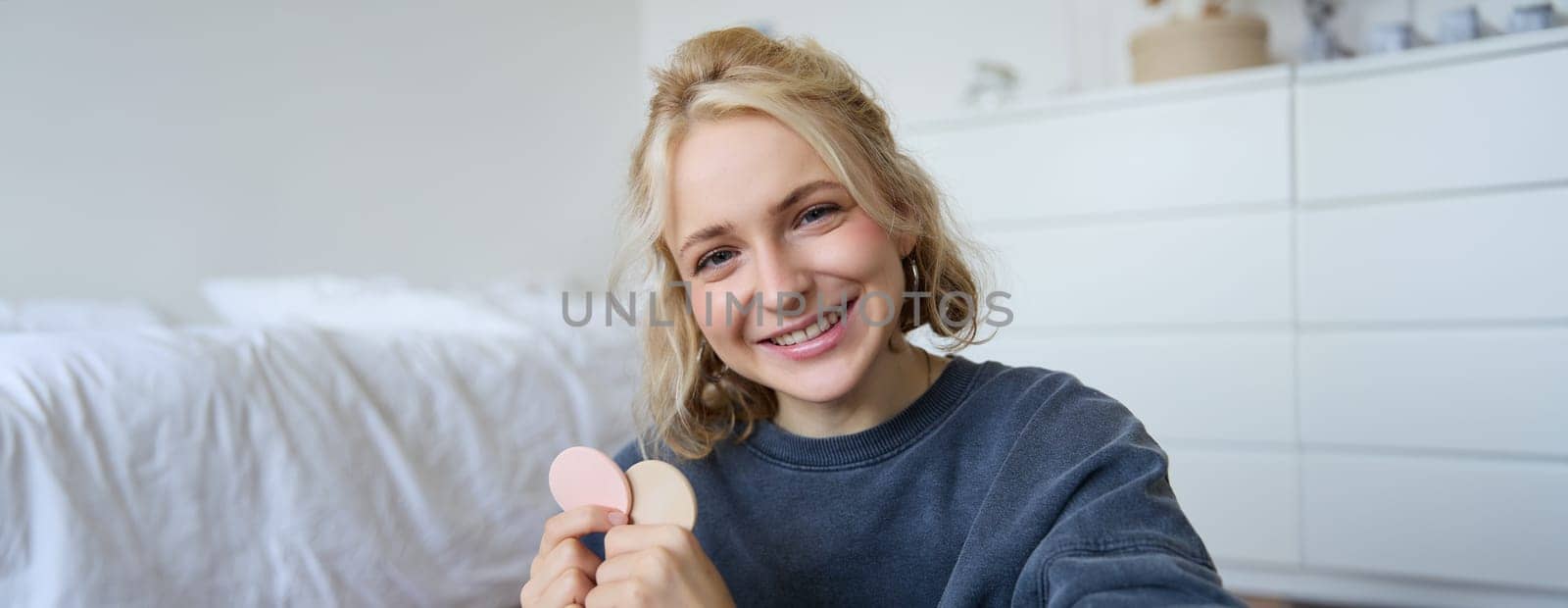 Close up portrait of happy, beautiful young vlogger, content maker recording video about makeup, showing beauty products on camera, smiling happily.