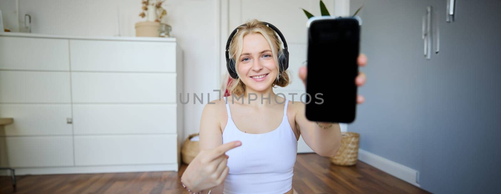Portrait of fitness instructor showing her favourite workout app, pointing finger at smartphone screen, showing mobile app on camera, smiling and looking satisfied, wearing headphones.