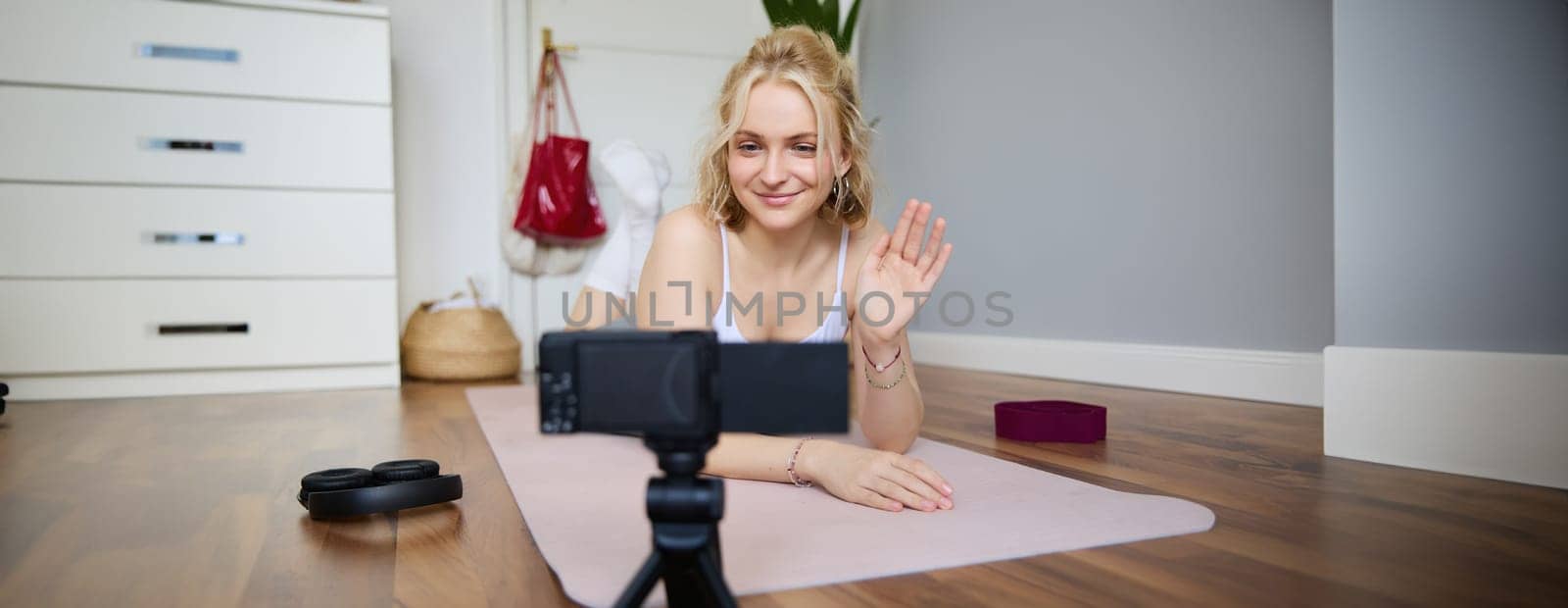 Portrait of smiling fitness instructor, social media content creator recording a vlog about sport, using rubber yoga mat, waving hand and looking at digital video camera by Benzoix