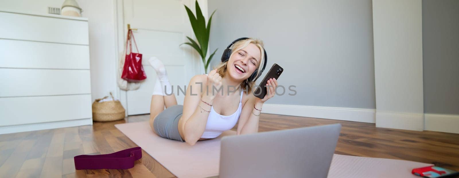 Portrait of woman lying on rubber yoga mat in room, wearing headphones, listens to music, using laptop and smartphone. Lifestyle and wellbeing concept