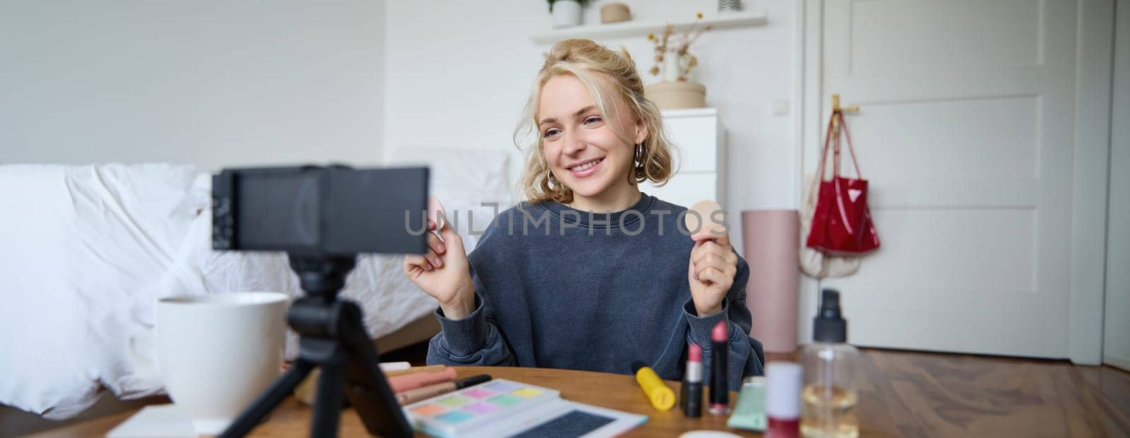 Portrait of young woman, beauty content creator, sitting in a room in front of digital camera, recording makeup tutorial vlog, showing cosmetic facial products.