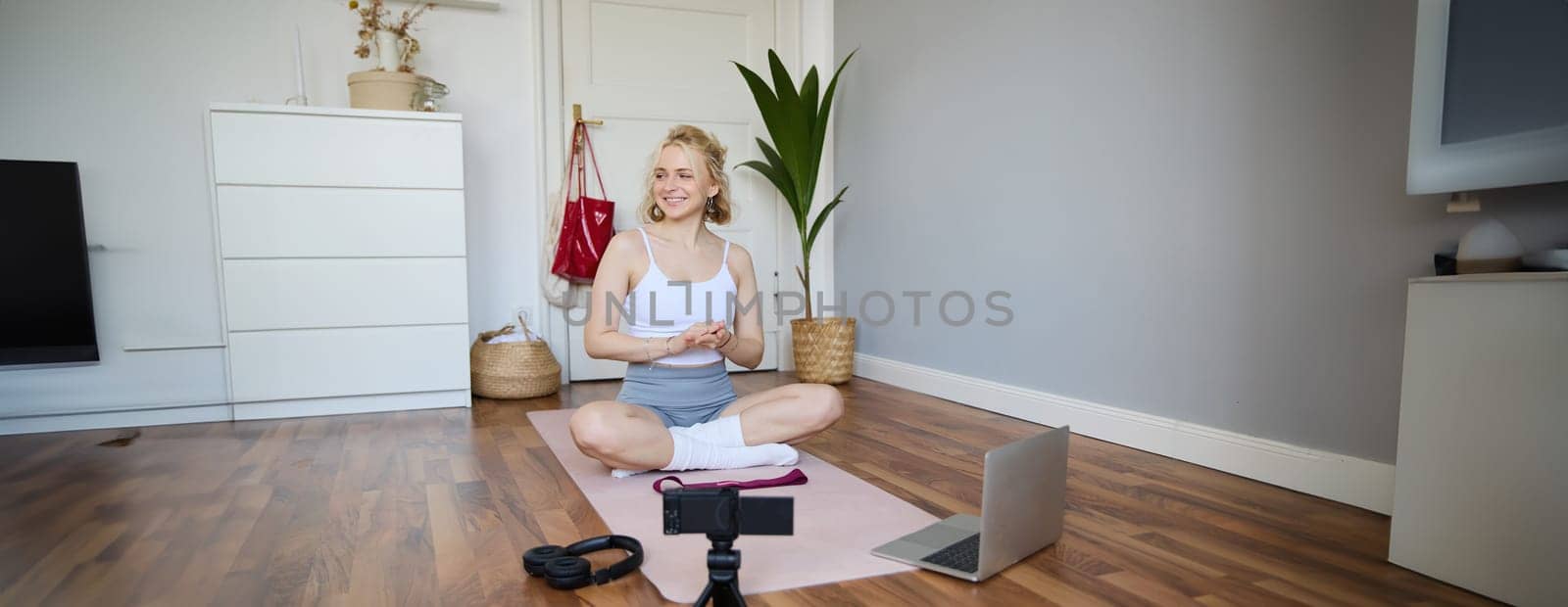 Portrait of young fitness blogger, woman showing exercises to her followers, recording video on digital camera, doing workout training session, home yoga, sitting in front of laptop on rubber mat by Benzoix