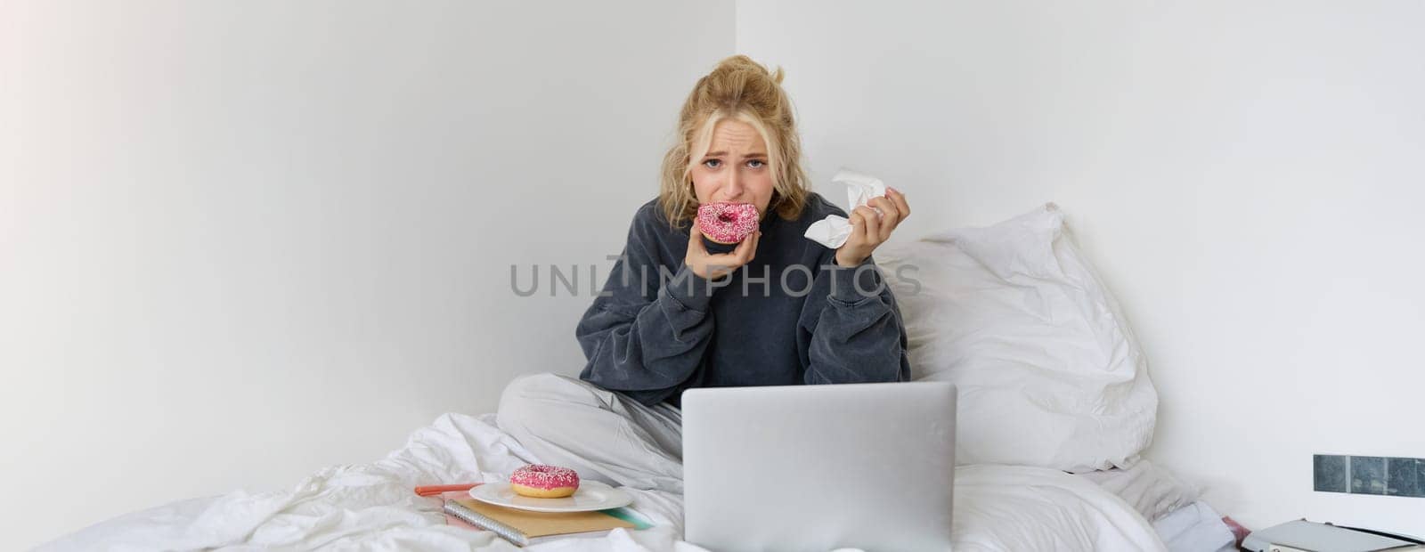Portrait of sad woman crying, eating doughnut, wiping tears off, looking at something upsetting on laptop screen, sitting on a bed.