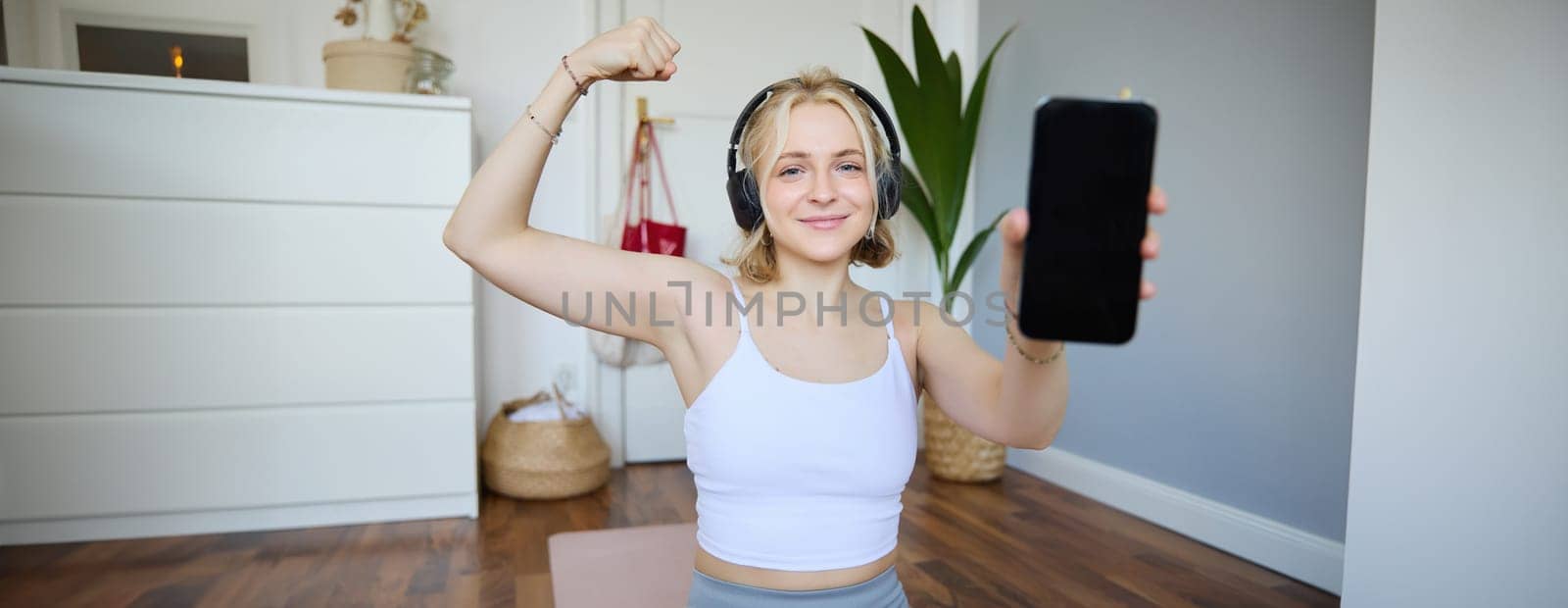Smiling blond woman working out, showing biceps, recommending smartphone app, demonstrating mobile phone screen at camera, sitting on rubber yoga mat in a room.