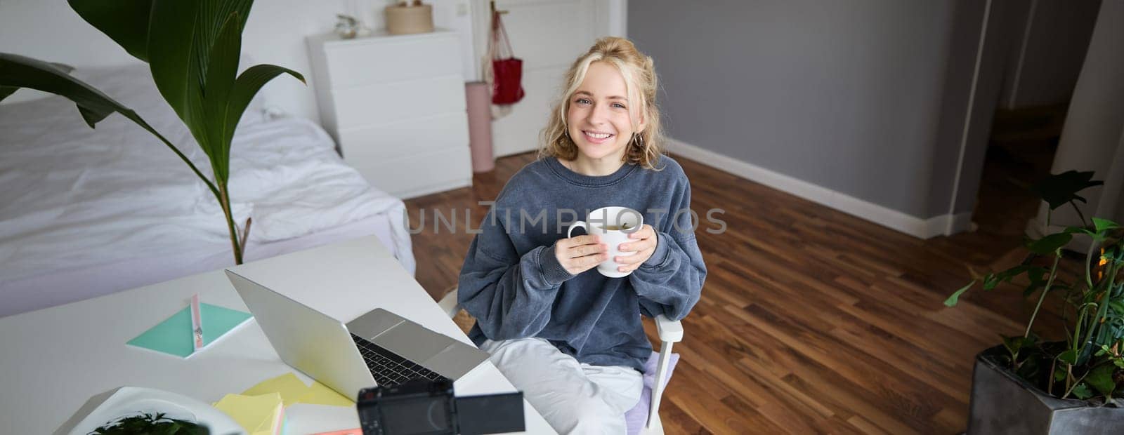 Image of young woman, social media influencer, editing her video on laptop, sits in a room with computer and digital camera, drinking coffee, smiling at camera.