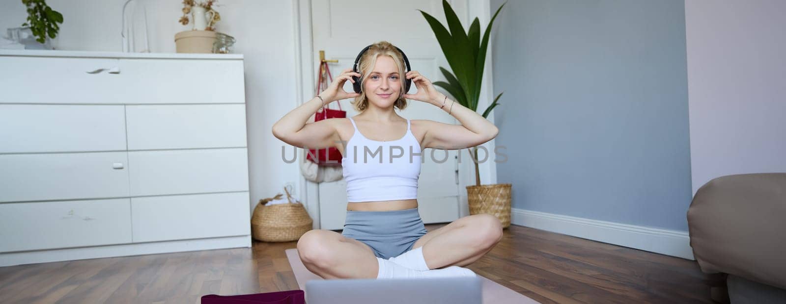 Portrait of young fitness girl, woman workout at home, listens to fitness instructor in wireless headphones, follows exercises on laptop.