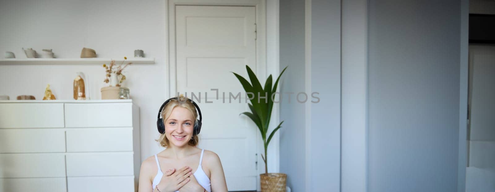 Portrait of woman feeling relaxed and in peace after meditation or yoga training at home, holding hands on chest, wearing headphones, smiling at camera by Benzoix