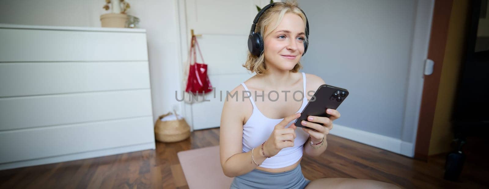 Portrait of young blond woman in headphones, turning on yoga, workout app on smartphone, choosing music on mobile phone application, sitting on rubber mat in room by Benzoix