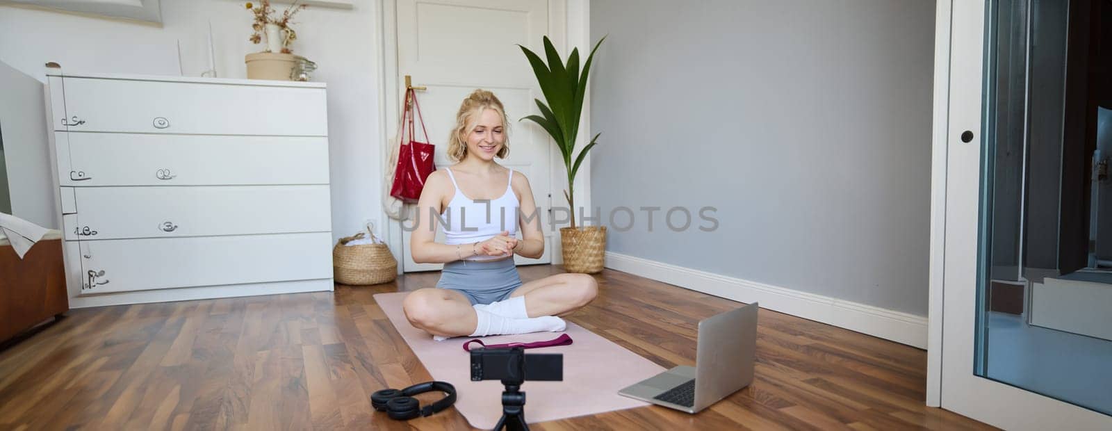 Portrait of young fitness blogger, woman showing exercises to her followers, recording video on digital camera, doing workout training session, home yoga, sitting in front of laptop on rubber mat.