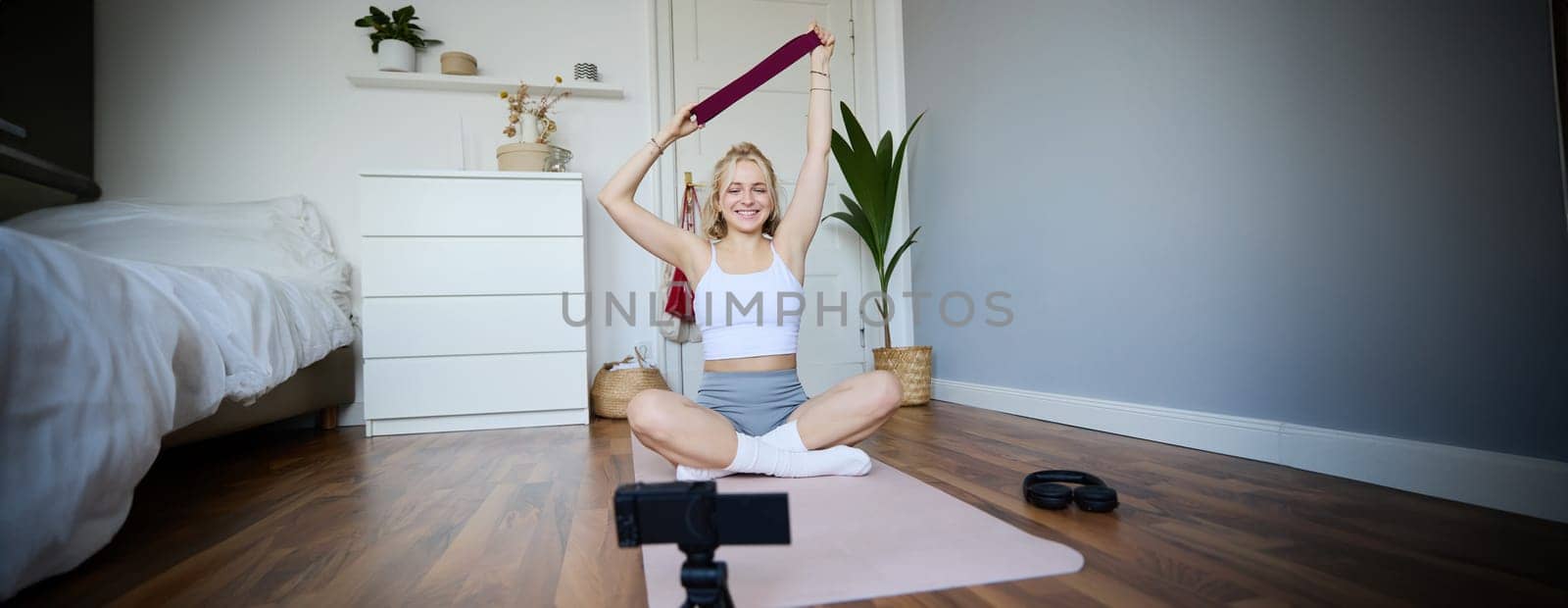 Smiling young blond woman recording video about workout, shooting on digital camera, fitness instructor shows how to use resistance band during exercises by Benzoix