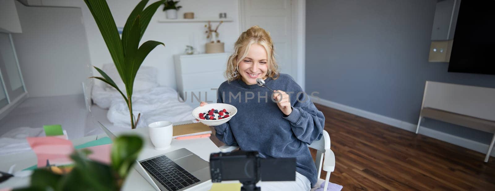 Portrait of smiling, candid young woman, content creator, eating bowl of dessert and looking at digital camera, recording vlog for followers on social media.