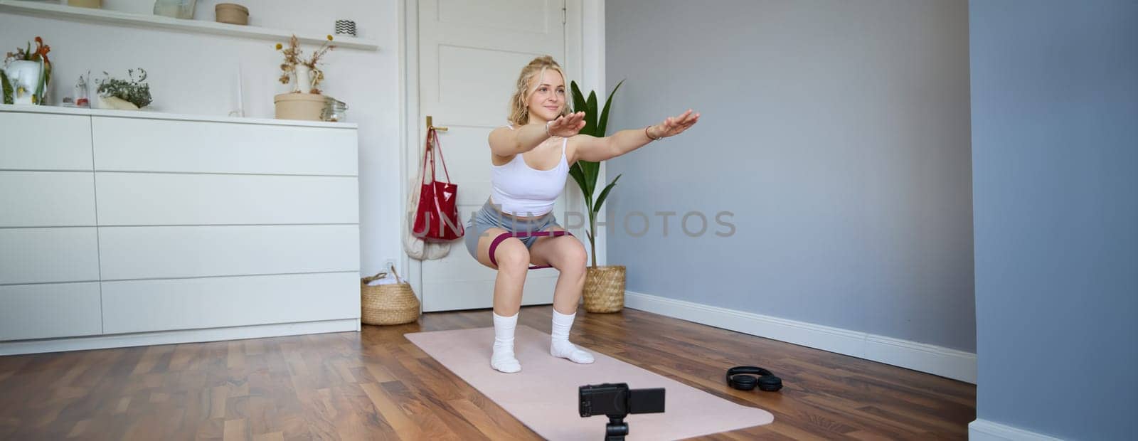 Portrait of woman, fitness instructor at home, recording video about workout, showing how to do leg exercises, squats with elastic resistance band, working out indoors on yoga mat by Benzoix