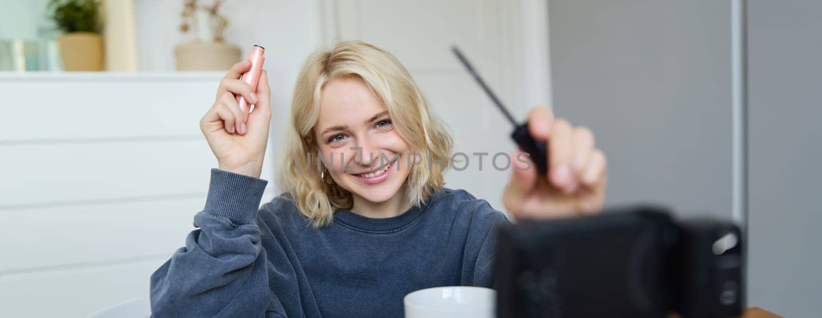 Portrait of young smiling woman in her room, recording video on camera, lifestyle vlog for social media, holding mascara, reviewing her makeup beauty products, showing how to use cosmetics by Benzoix