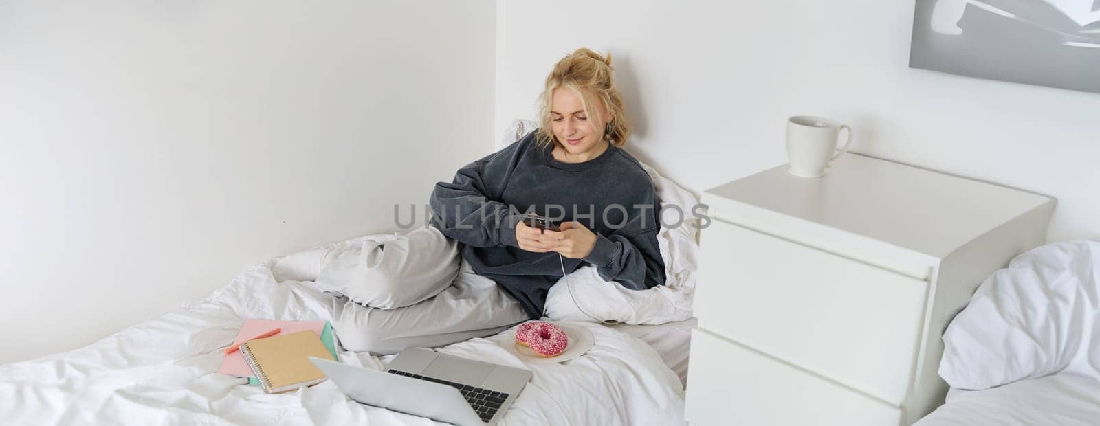 Portrait of smiling candid woman, lying in bed with doughnut, using smartphone and laptop, resting at home in bedroom, watching tv show or chatting online.