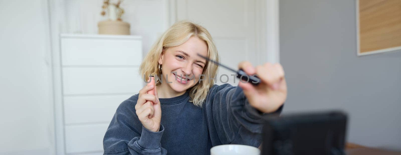 Portrait of young smiling woman in her room, recording video on camera, lifestyle vlog for social media, holding mascara, reviewing her makeup beauty products, showing how to use cosmetics.