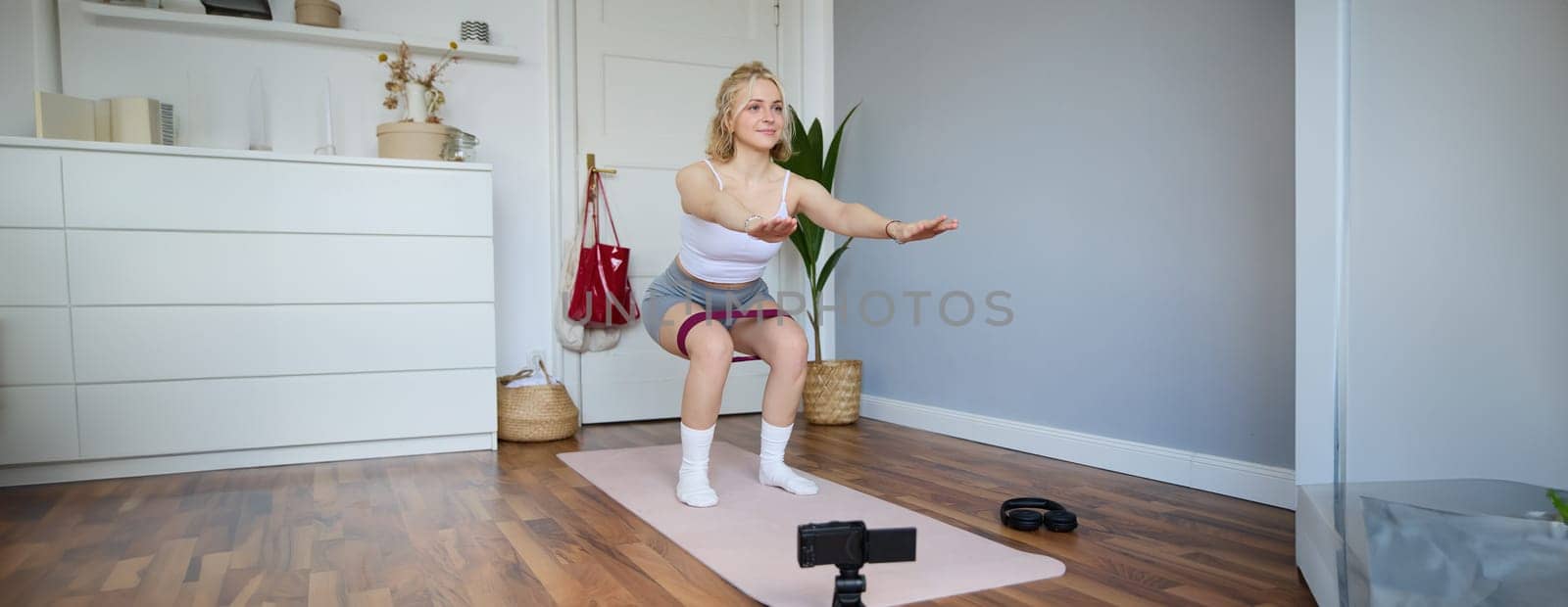 Portrait of young woman, fitness instructor making a video for sport vlog, doing squats on camera, using resistance band, working out at home.
