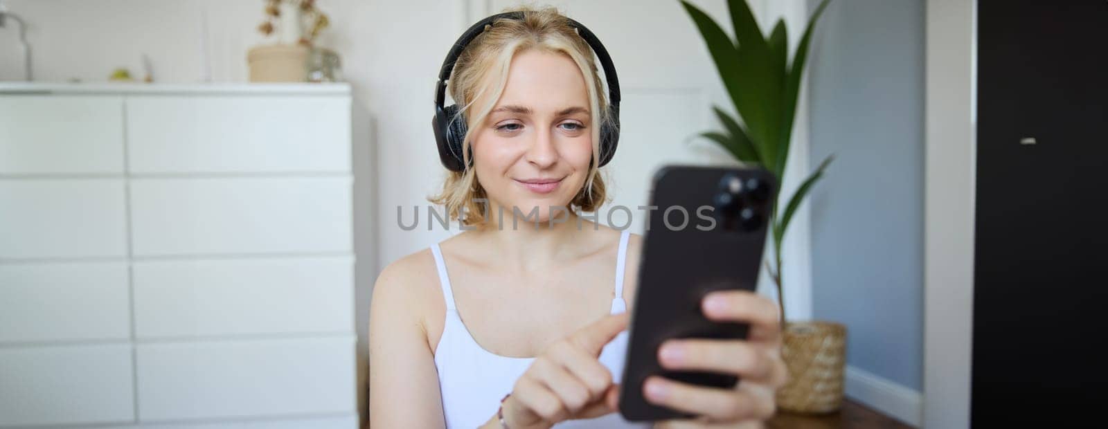 Close up portrait of smiling sporty woman, wearing headphones, using mobile phone, workout app while exercising at home. Wellbeing and lifestyle concept