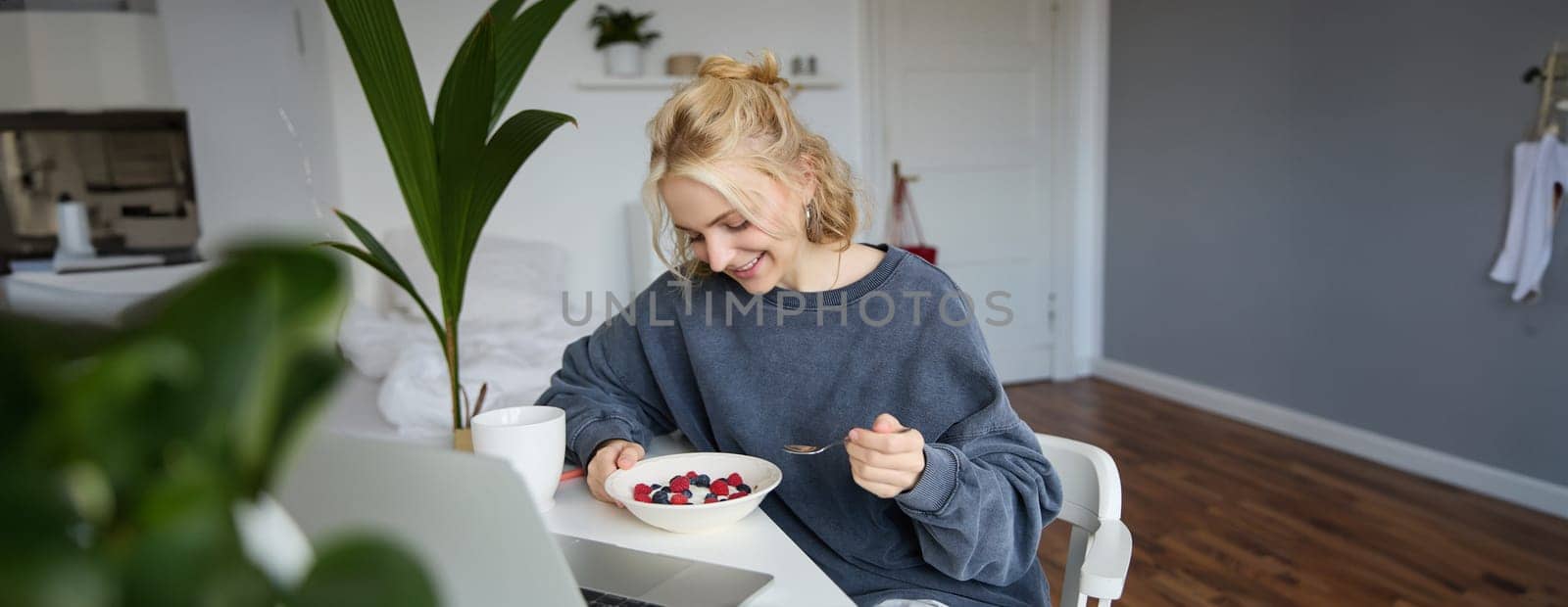 Portrait of smiling blond young woman, eating in front of laptop, watching videos online while having breakfast, enjoying dessert, sitting in bedroom by Benzoix