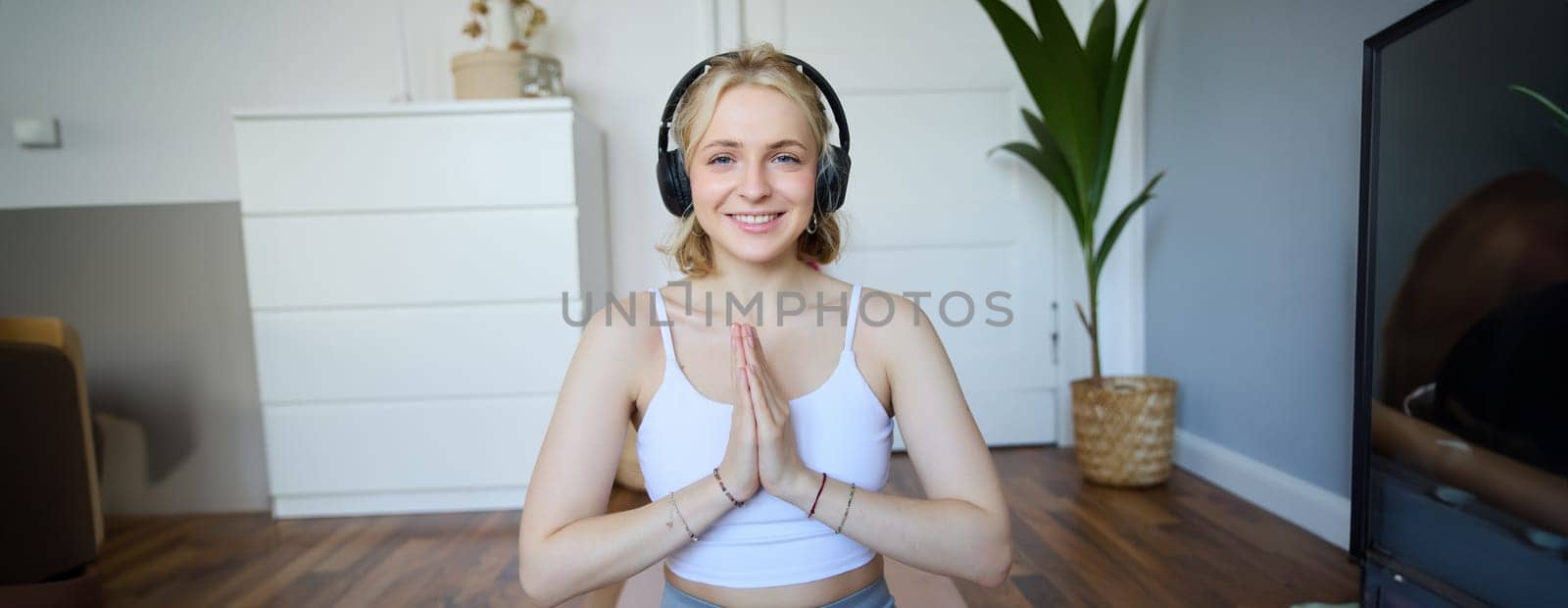 Portrait of blond young woman meditating, practise yoga, using yoga podcast to workout at home, sits on rubber mat by Benzoix