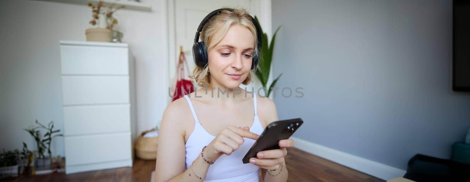 Portrait of young blond woman in headphones, turning on yoga, workout app on smartphone, choosing music on mobile phone application, sitting on rubber mat in room.