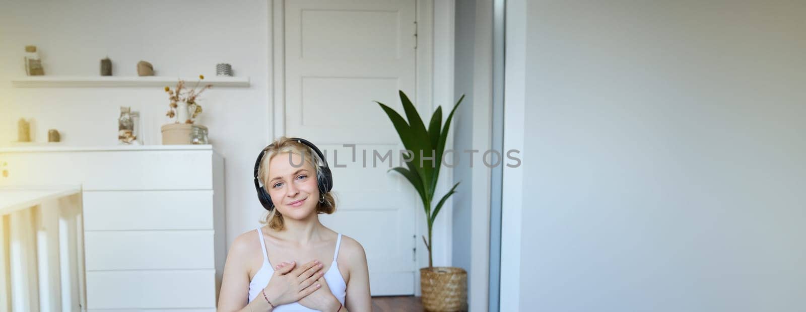 Vertical portrait of beautiful young woman in headphones, sits on yoga mat, meditating, holding hands on her chest and smiling at camera by Benzoix