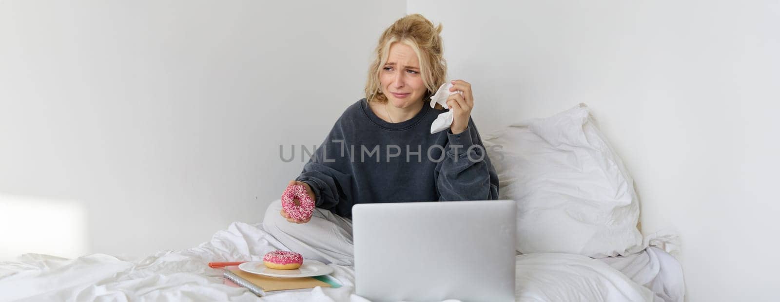 Portrait of woman sitting on a bed with laptop, eating doughnut and crying from sad movie scene.