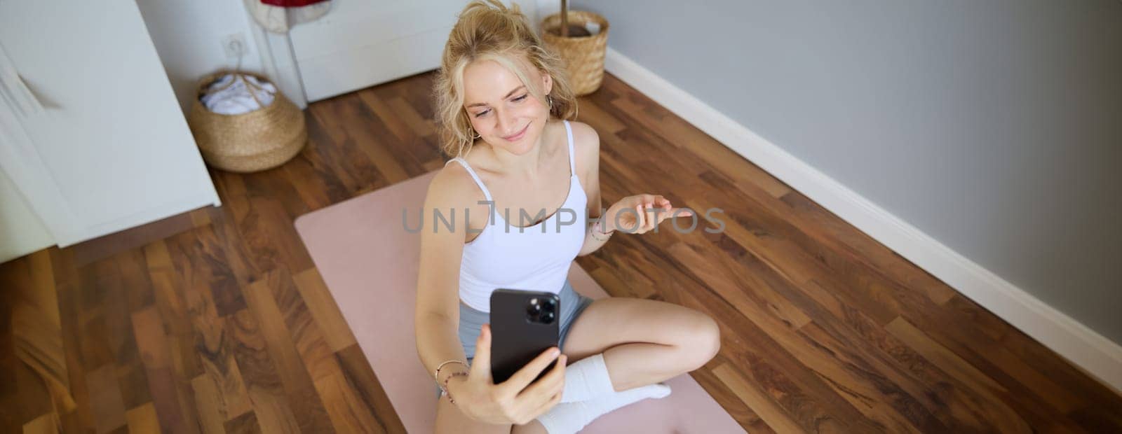 Portrait of woman having live stream training session, using her smartphone to create workout content, blogging during exercises or doing yoga, sitting on rubber mat at home by Benzoix