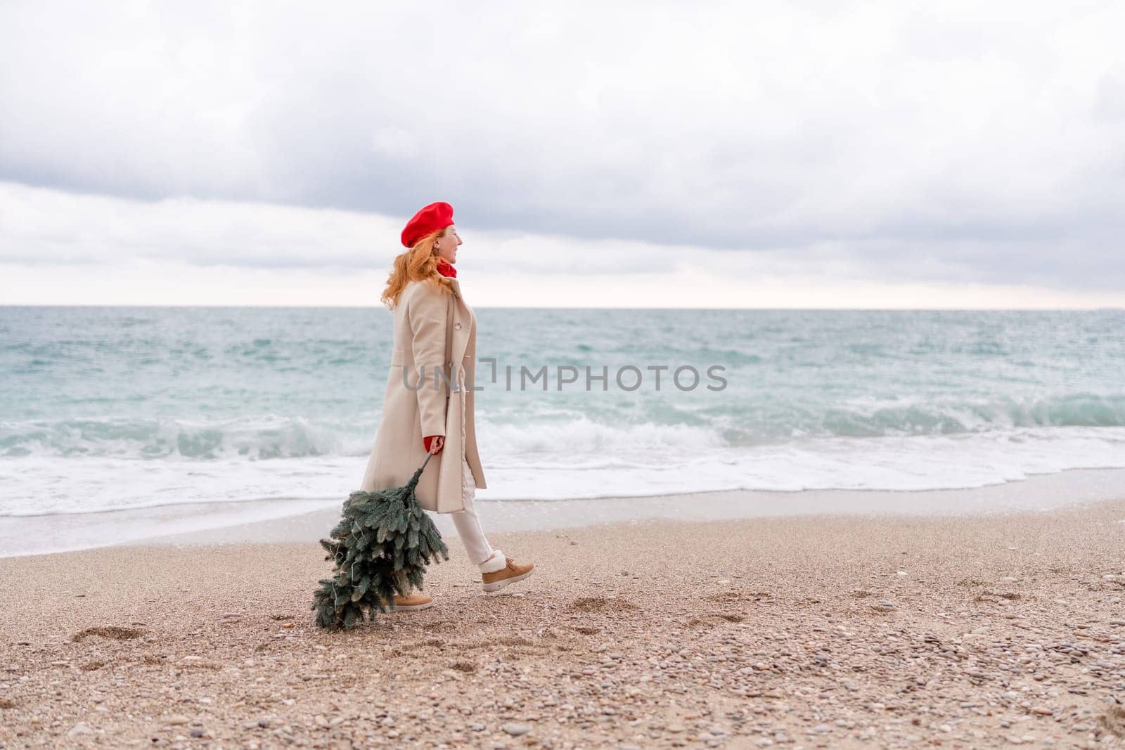 Redhead woman Christmas tree sea. Christmas portrait of a happy redhead woman walking along the beach and holding a Christmas tree in her hands. She is dressed in a light coat and a red beret. by Matiunina