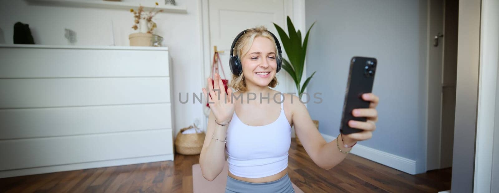 Portrait of young sporty woman in headphones, records video on her smartphone, live streaming and saying hello to followers while doing workout training at home by Benzoix