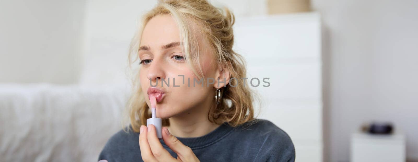 Close up portrait of beautiful blond woman applies lip gloss on her lips, puts on makeup, gets ready for night out, using beauty products, looks in mirror by Benzoix