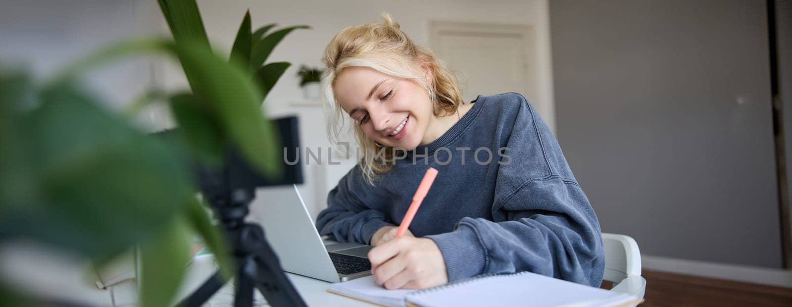 Portrait of young woman, lifestyle blogger, recording video of herself, making notes, writing in journal, sitting in front of laptop in a room and studying.