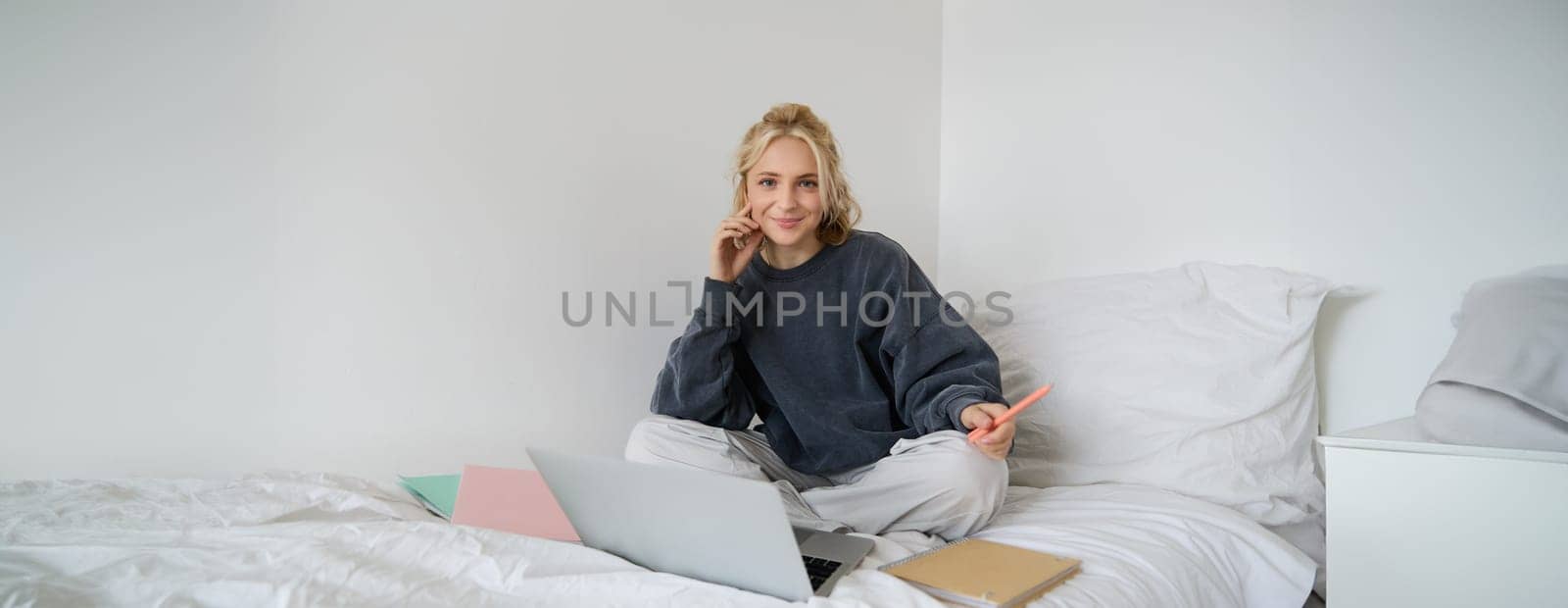 Portrait of young smiling woman studying in her bed, working from home in bedroom, sitting with laptop and notebooks on lotus pose, looking happy and relaxed.