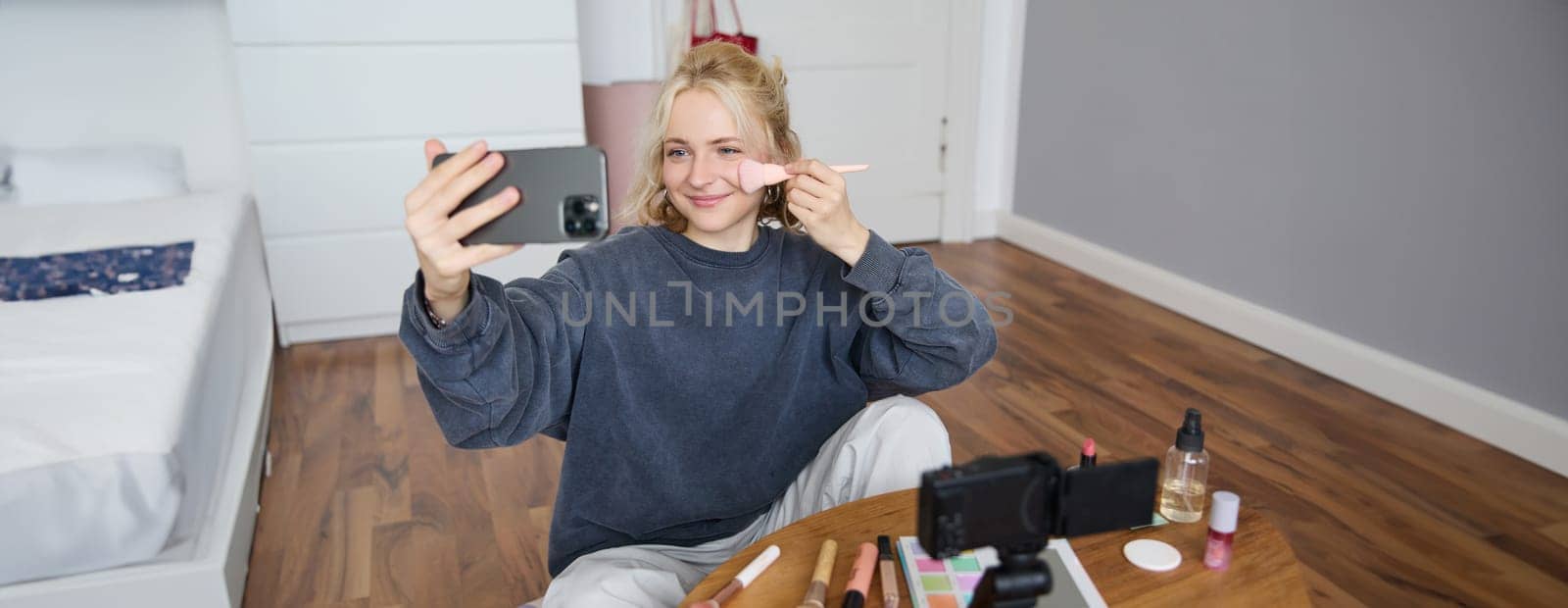 Portrait of beautiful blond girl doing makeup on camera, recording vlog using digital camera and live stream on mobile phone app, chatting with audience.