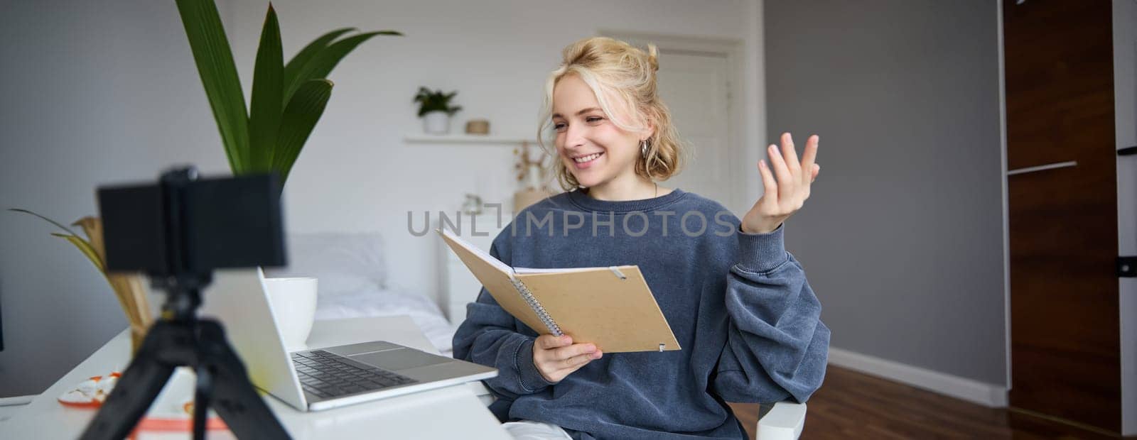 Image of young blong woman, student studying online at home, sits in her room, reads notes, chats via laptop. attends course, works on remote.