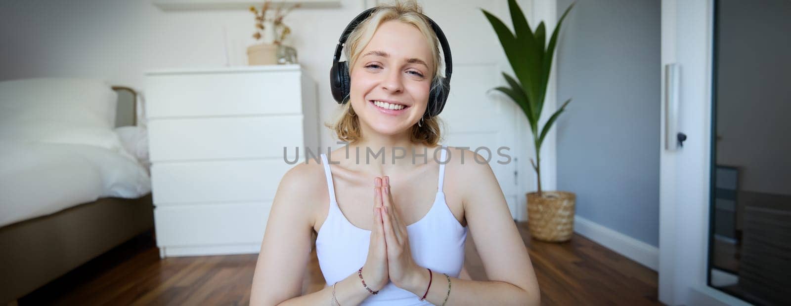 Portrait of woman meditating at home, sitting in headphones, listening to yoga podcast, holding hands in namaste sign by Benzoix