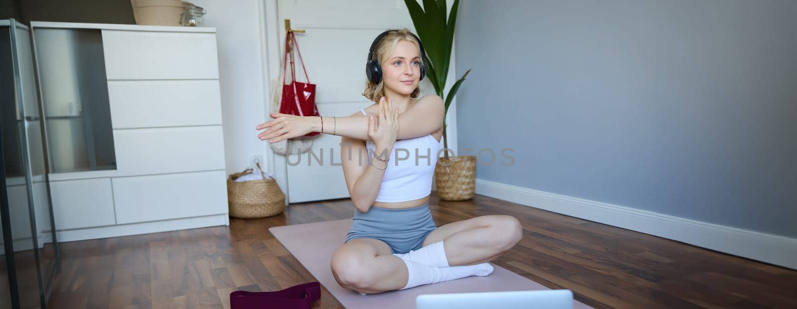 Portrait of beautiful, athletic woman at home, doing workout, watching fitness instructor video on laptop, wearing headphones, stretching her arms.