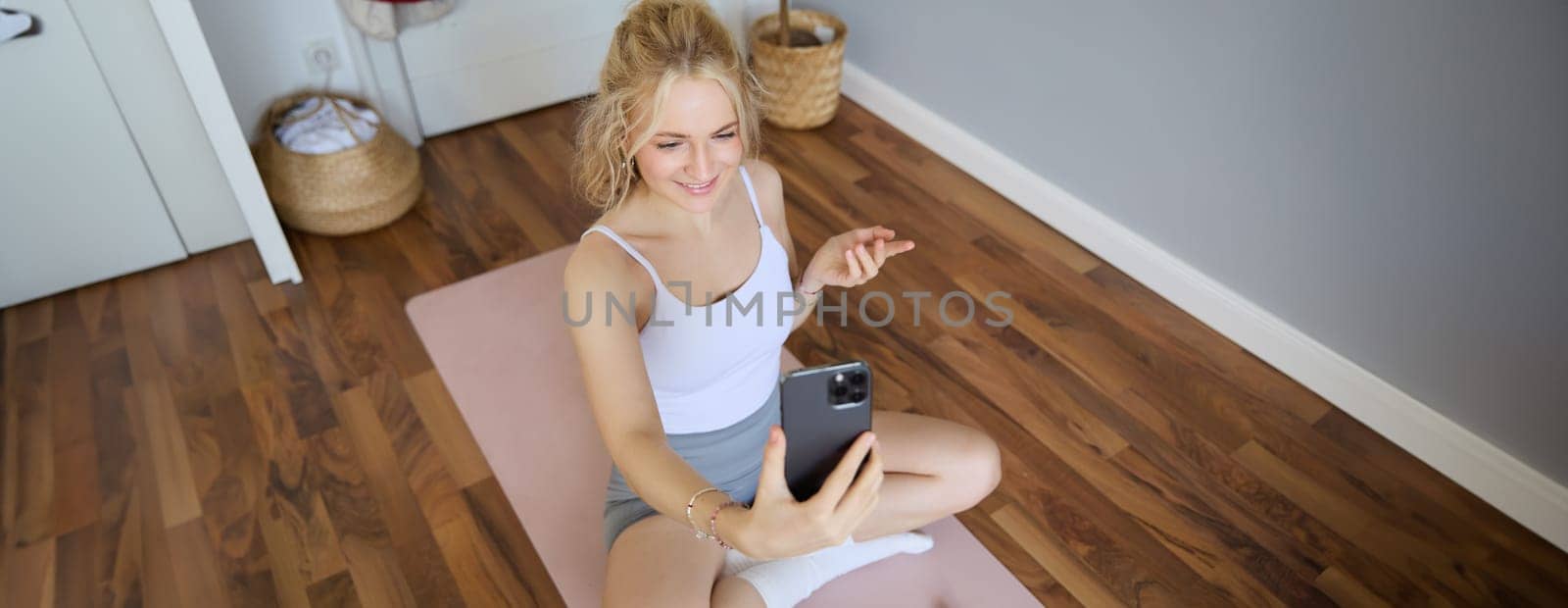 Portrait of young blogger, fitness instructor showing how to do exercises, sitting on yoga mat with smartphone, talking to followers on live stream.