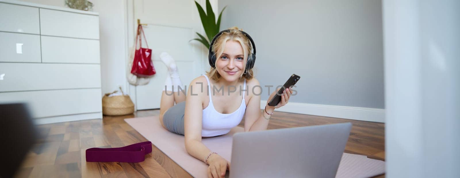 Portrait of young sporty woman, connects to online workout training session, doing exercises, lying on rubber mat, listens to fitness instructor in headphones, holding mobile phone by Benzoix