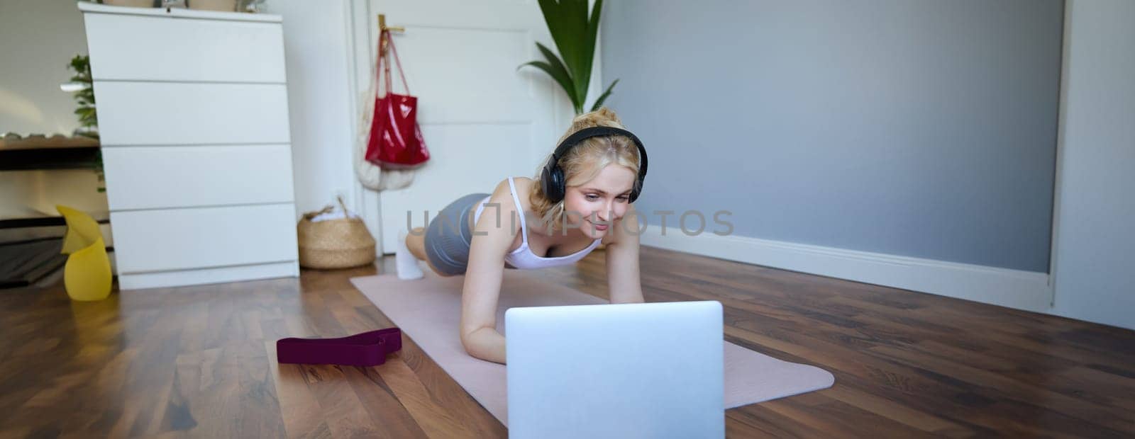 Portrait of young sporty woman doing workout, looking at fitness video on laptop in wireless headphones, standing in plank on yoga mat, following exercise tutorial by Benzoix