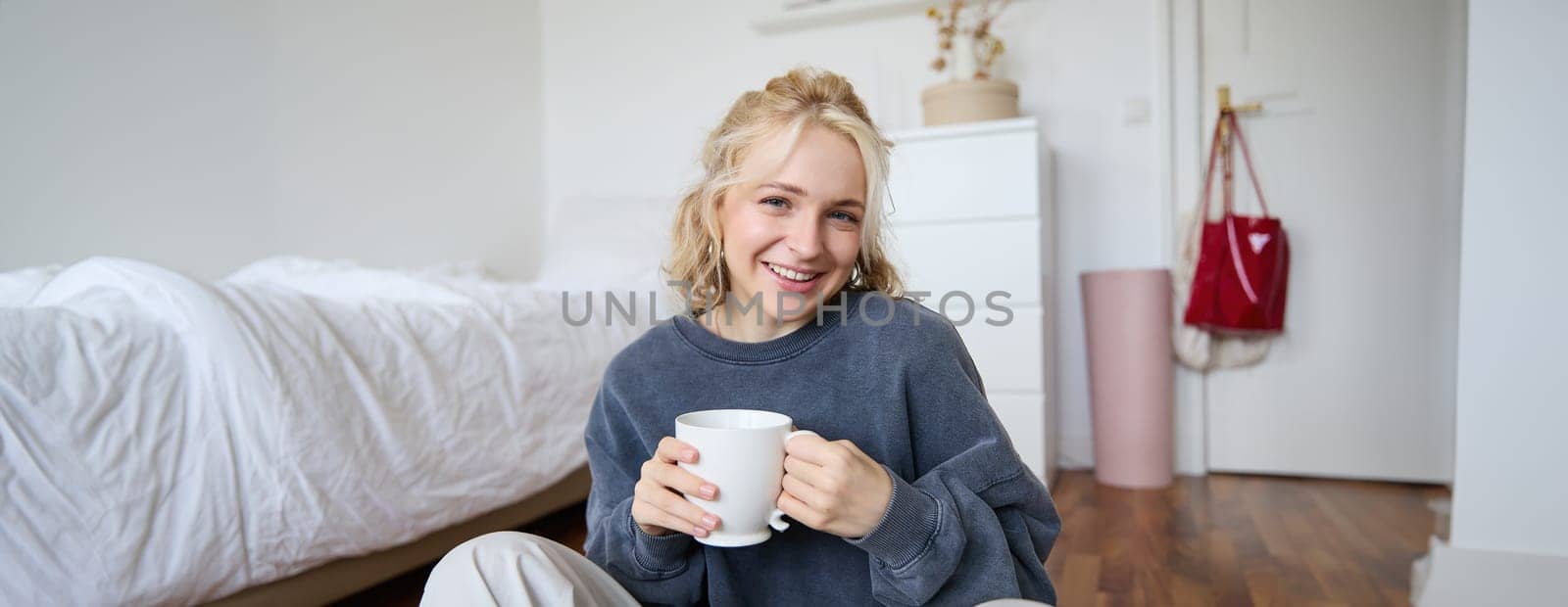 Portrait of young beautiful woman in casual clothes, sitting on bedroom floor with cup of tea, drinking and smiling, looking at camera, recording a lifestyle vlog.